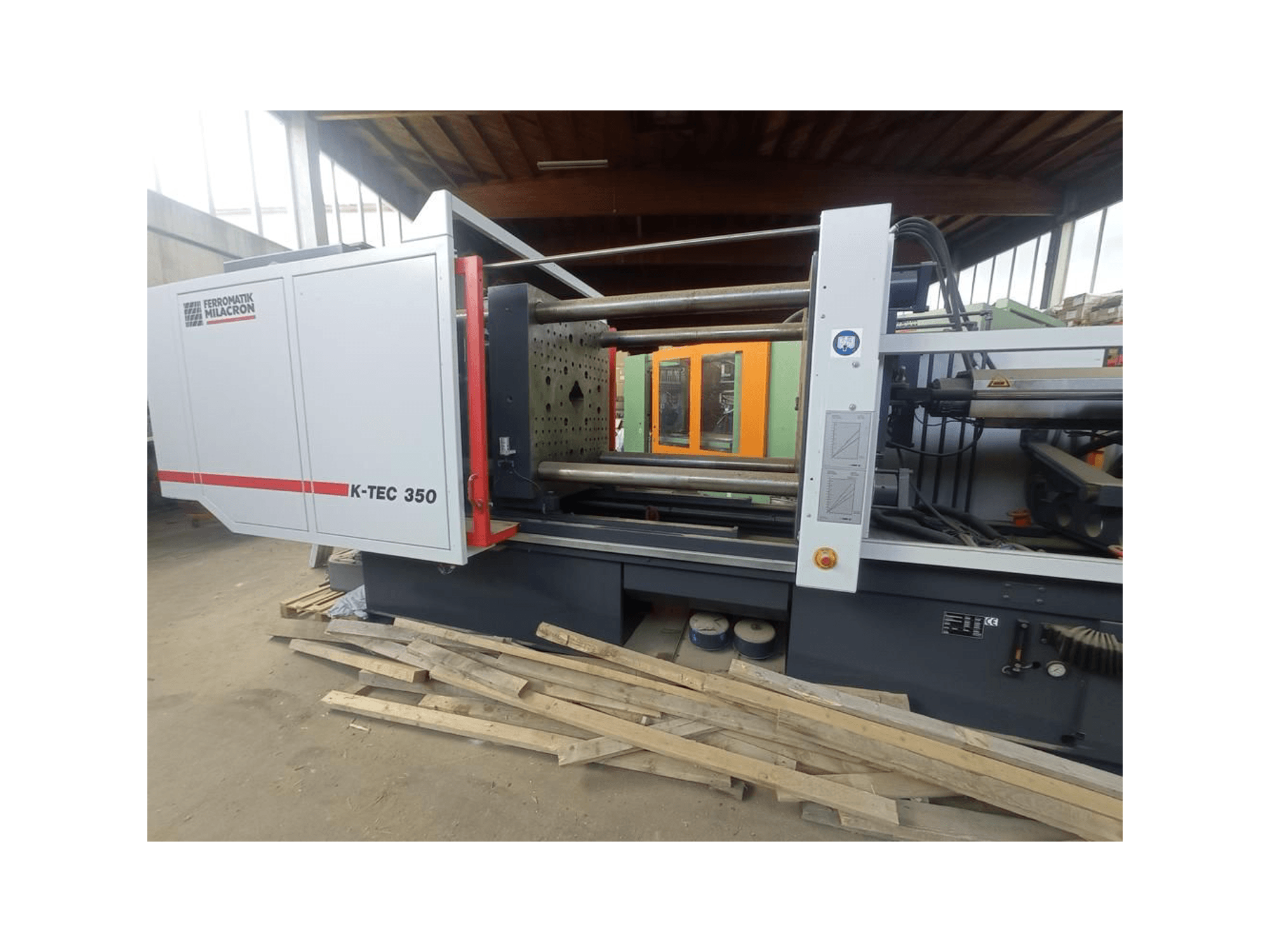 Ferromatik Milacron K-TEC 350, skatoties no sāniem, ar sarežģītiem mehānismiem un darbības vadības ierīcēm.