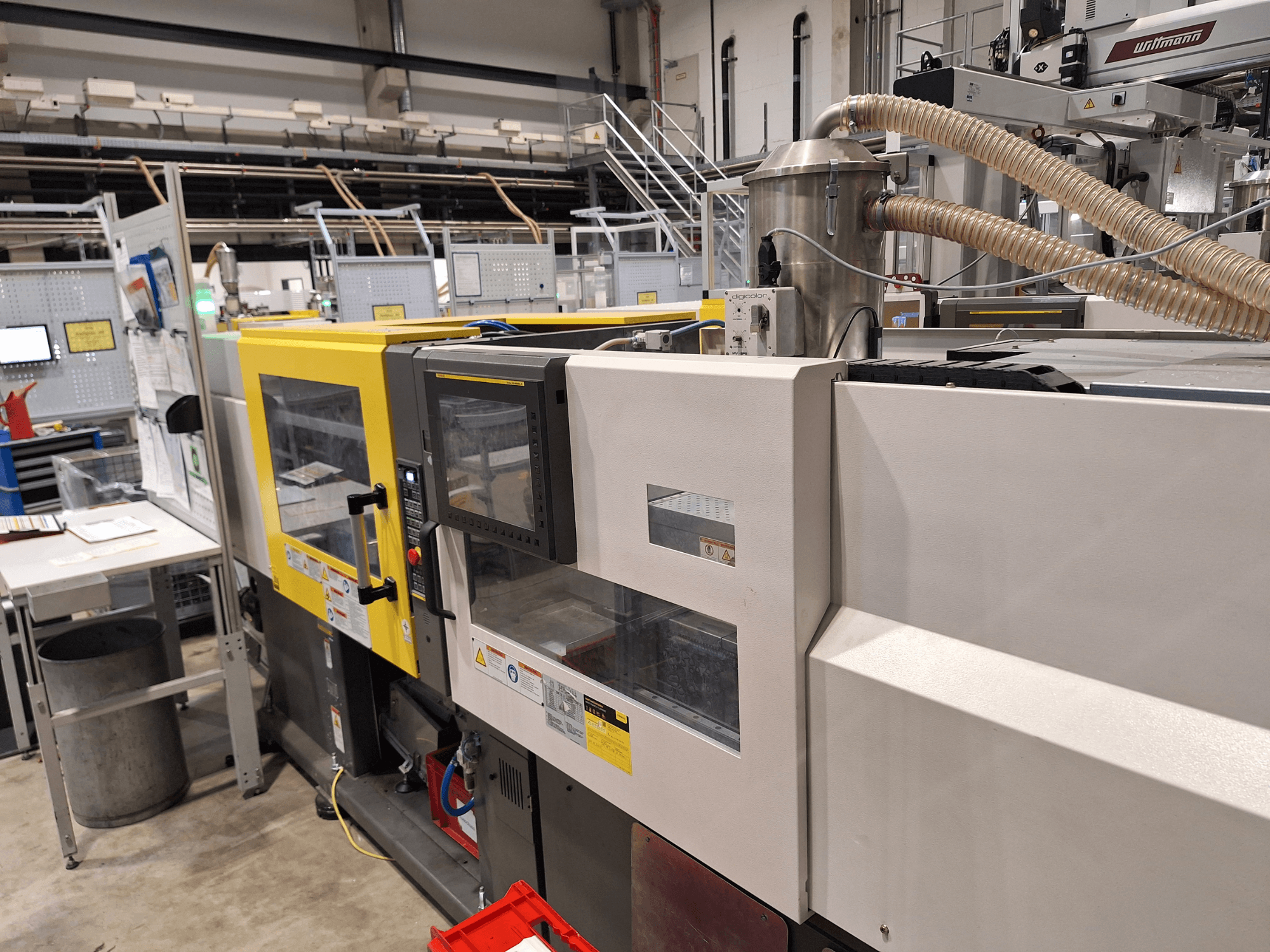 Skats no priekšpuses uz daudzkrāsainu injekcijas formēšanas iekārtu ar dzeltenām un pelēkām sekcijām, ar vadības paneli un izplūdes sistēmu.