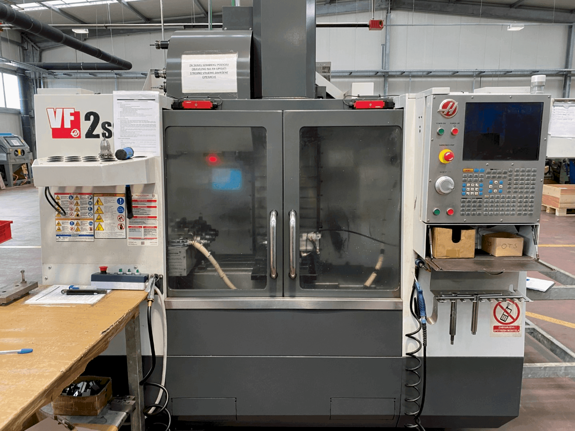 Haas VF-2SS apstrādes centrs priekšpusē, ar vadības paneli, dzesēšanas šķidruma šļūteni un drošības marķējumu sānos.